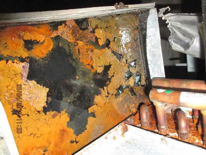 A corroded drainage tray, orange rust across the metal beside copper pipework