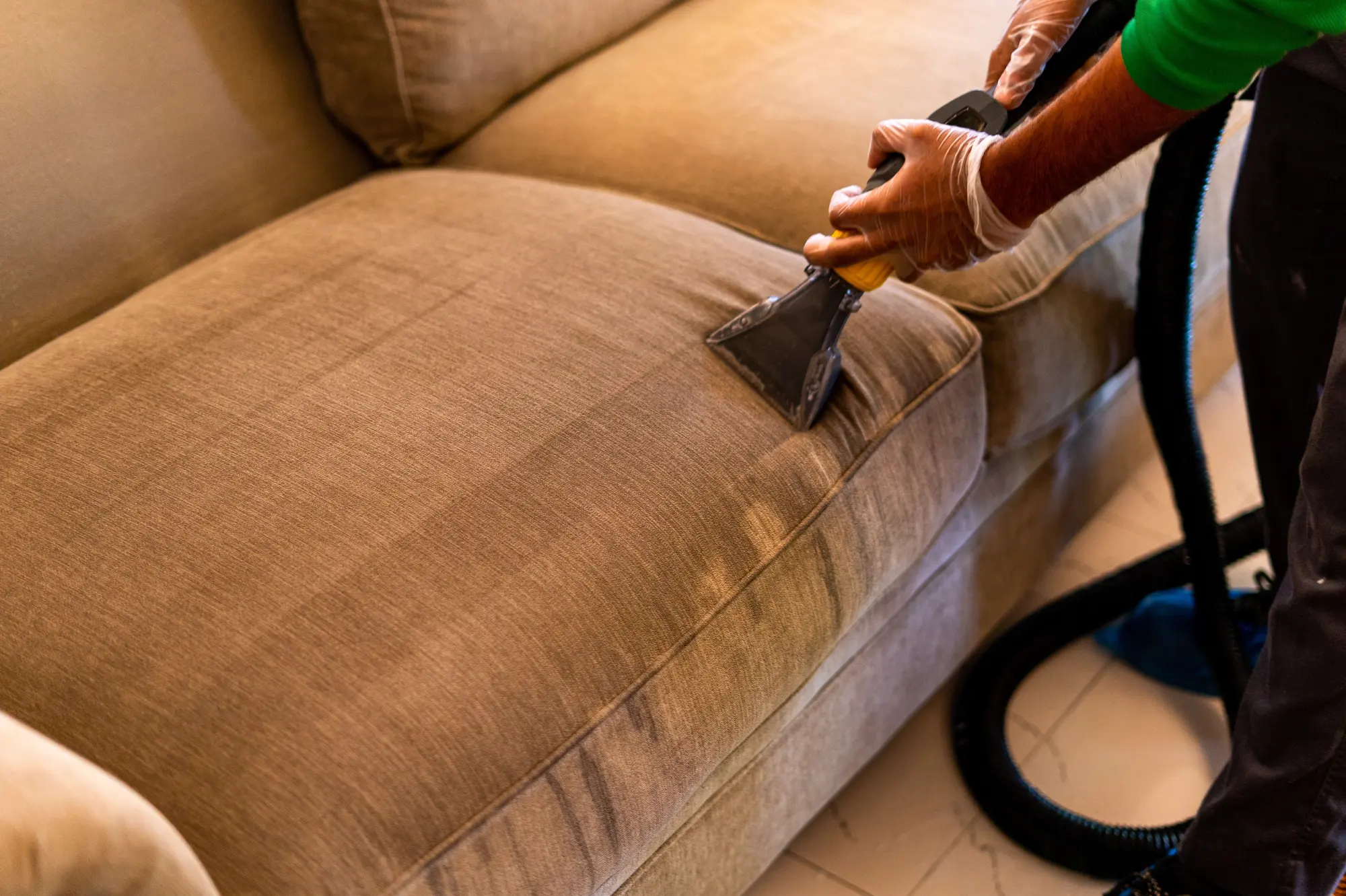 A gloved hand drawing an extraction tool across a brown fabric sofa, the cleaned stripe visible behind it