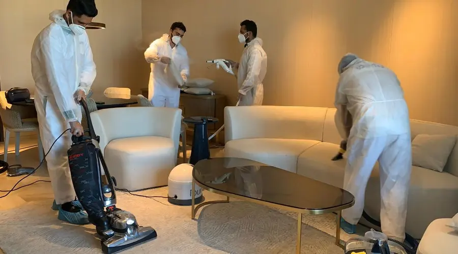 Four iPurity technicians in white protective suits sanitising a living room, one vacuuming the carpet while the others treat the furniture